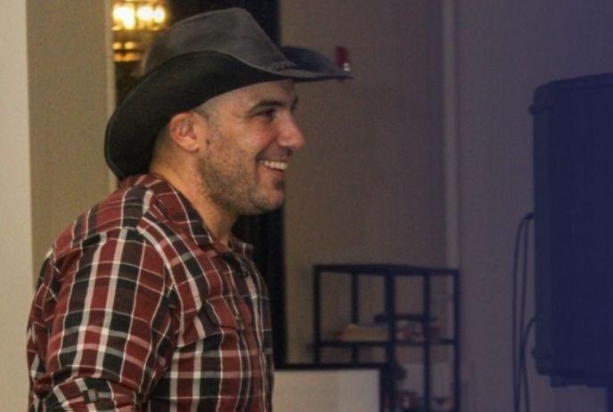 Man in cowboy hat and plaid shirt dances indoors; crowd watches under warm lights.