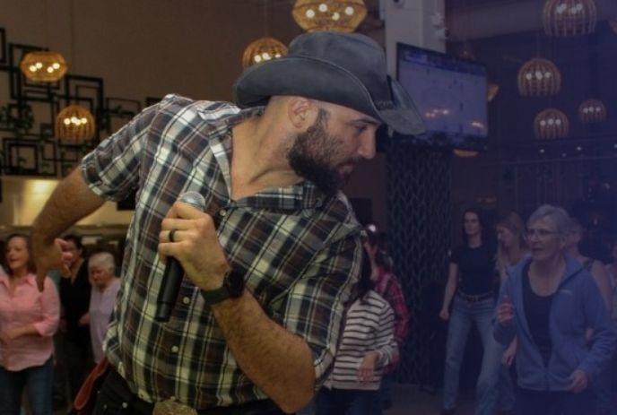 Man in cowboy hat and plaid shirt holds mic mid-dance, warm indoor crowd watches under hanging lights.