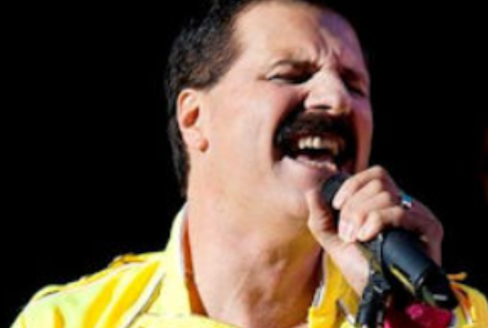 Person dressed as Freddie Mercury with yellow shirt and jacket singing into a microphone.