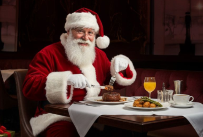 Santa at a table holding a knife and fork eating a full meal.