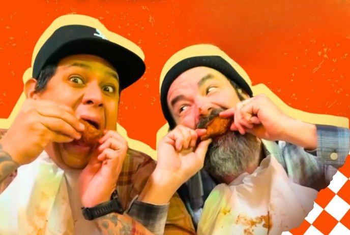 Two men enjoying chicken wings amid orange backdrop, one tattooed, one bearded in beanie.