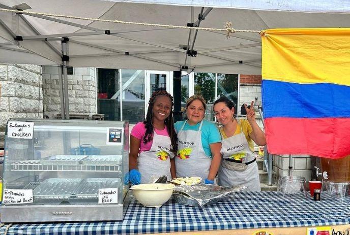 Three vendors in aprons serve empanadas at an outdoor stall beside a bright Colombian flag.
