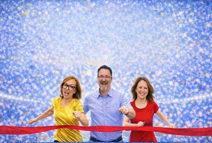 Three comedians, 2 women in a red and a yellow top and a man in blue shirt crossing a red ribbon, finishing a race.