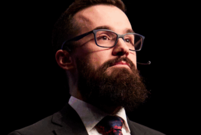 Keith Brown wearing glasses and a shirt & tie, looks ahead for a photo.