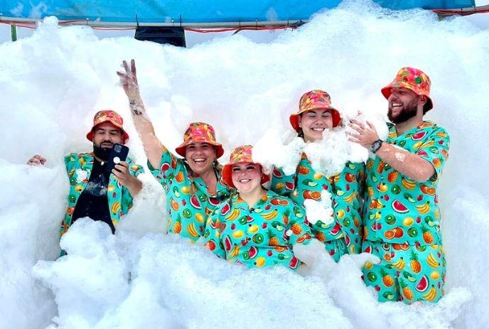 Five individuals in fruit-print outfits enjoy a foam party, smiling and taking selfies in thick white bubbles.
