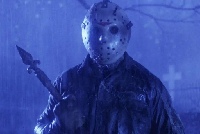 Masked figure stands in rain holding weapon before grave in dark blue night scene.