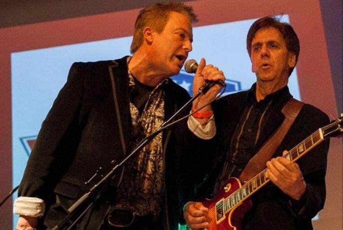 Two musicians perform onstage one singing and one playing guitar under bright event lighting.