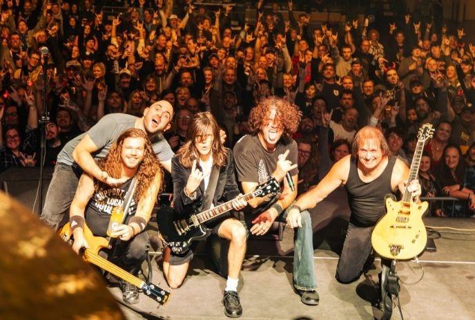 Five band members kneel onstage before a cheering crowd holding up glowing phones in a packed theater