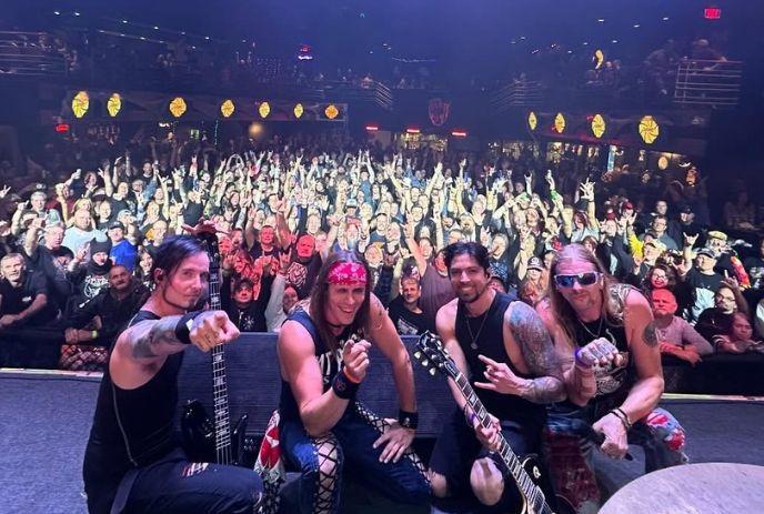 Four band members kneel at the stage edge as a packed crowd cheers behind them under the spotlights.