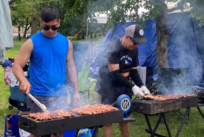 Two people grilling skewered meat on large charcoal grills in a park with smoke rising around them