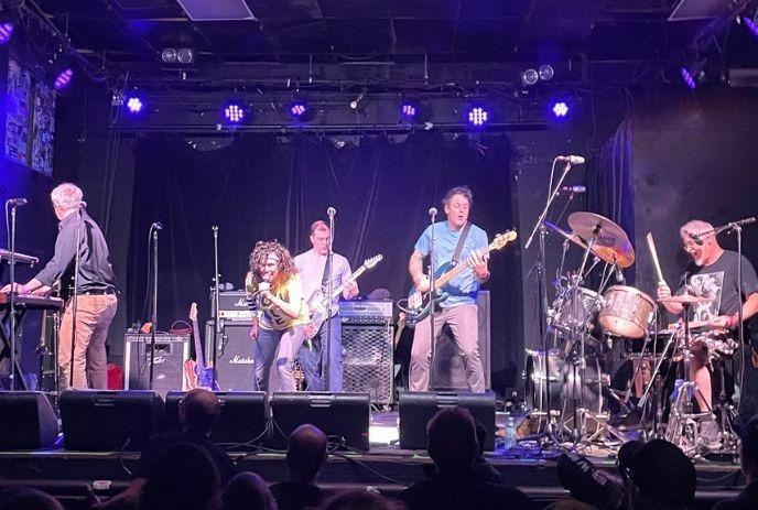 A band playing on the stage under the blue light and crowd cheering.