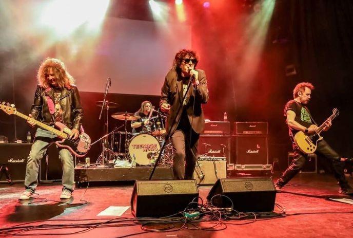 Rock band performing on stage with singer guitarist bassist drummer and bright colored lights.