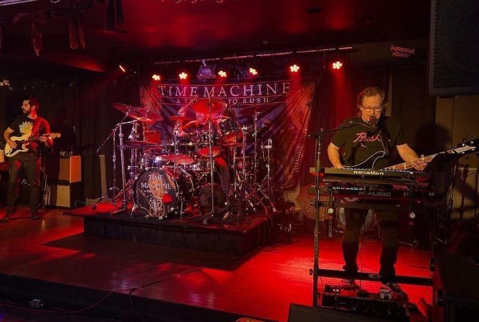 Tribute band performing on stage with drummer guitarist and keyboardist under warm concert lights.