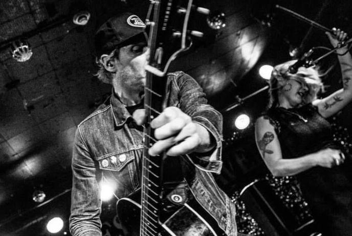 Guitarist in denim playing onstage with drummer behind him in a lively black and white frame.
