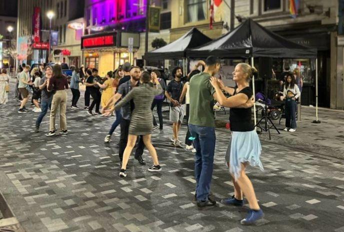 Night street dancers moving in pairs on patterned pavement with tents buildings and bright city lights.
