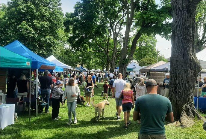 A lively crowd moves through shaded tents at a sunny park market lined with trees and vendors.