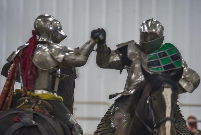 Two armored riders on horses meet in a friendly gesture inside an indoor arena setting.