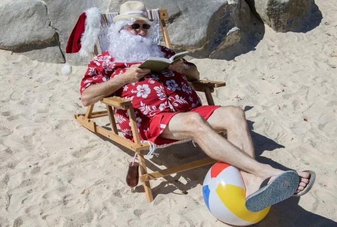 Santa in bright summer wear reads on a beach chair with sand rocks and a ball nearby.