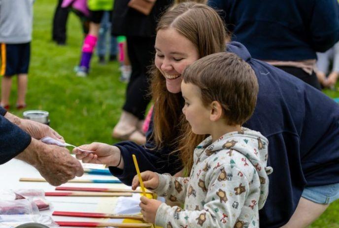 adult and child at table with colorful sticks outdoors engaging with others in friendly community event setting.