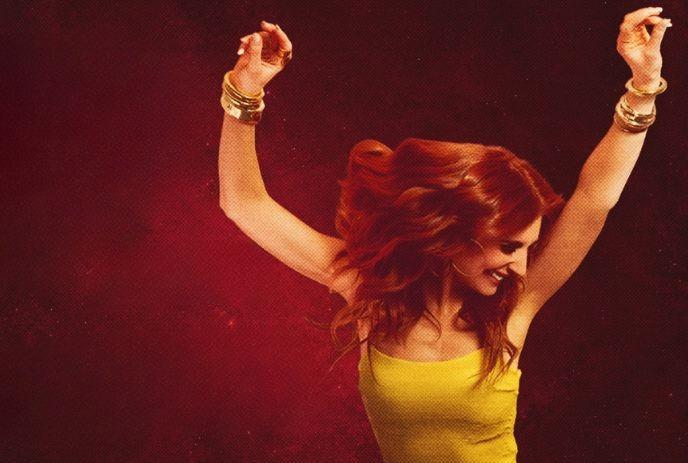 Artist with long wavy hair in yellow top arms raised against deep red backdrop suggesting lively motion.