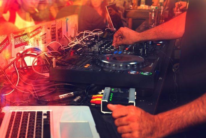 DJ works mixer and laptop amid colorful lights with crowd behind in a party vibe.