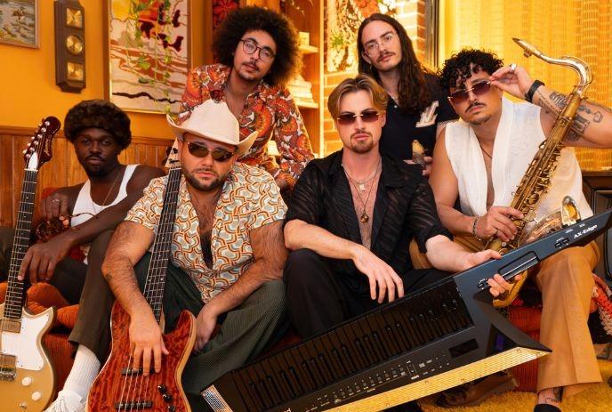 Six musicians in retro room with warm light holding instruments wearing vintage patterned outfits posing.