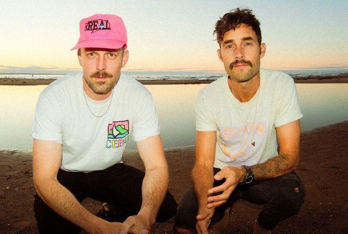 Artists kneeled on the beach at sunset wearing casual shirts and caps beside calm water in warm light