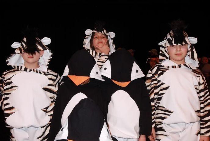 Children in zebra and penguin costumes stand together against dark backdrop creating playful lively scene.