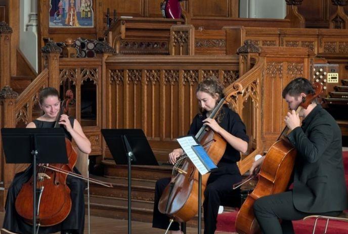Three musicians seated playing cellos in ornate church before carved wood and organ creating calm scene.