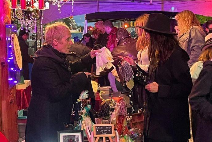 People browse warm lit market stall as two talk over displayed crafts beneath tent creating lively scene.