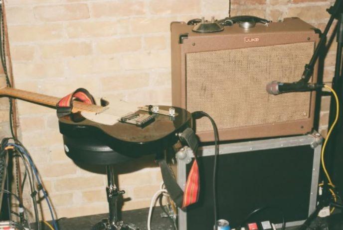 Electric guitar on stool beside amp and mic in brick walled space suggesting a ready music setup.