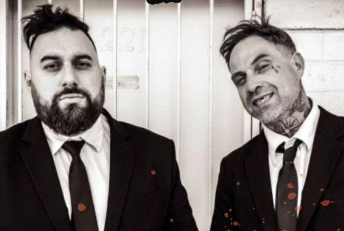 Two suited artists stand before a door in stark black, white style frame.