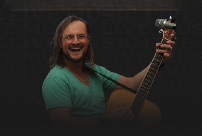 Smiling person in glasses holding acoustic guitar before dark backdrop with subtle live music theme.