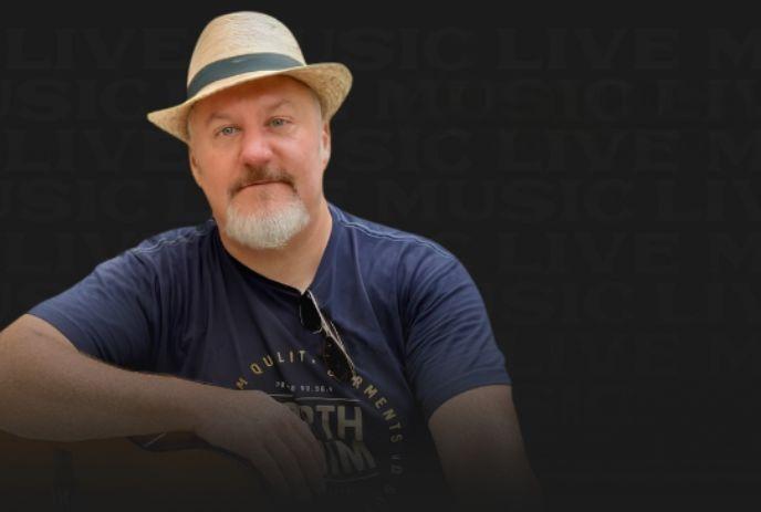 Artist in straw hat and dark shirt resting arm on guitar before dim backdrop with subtle live music theme.
