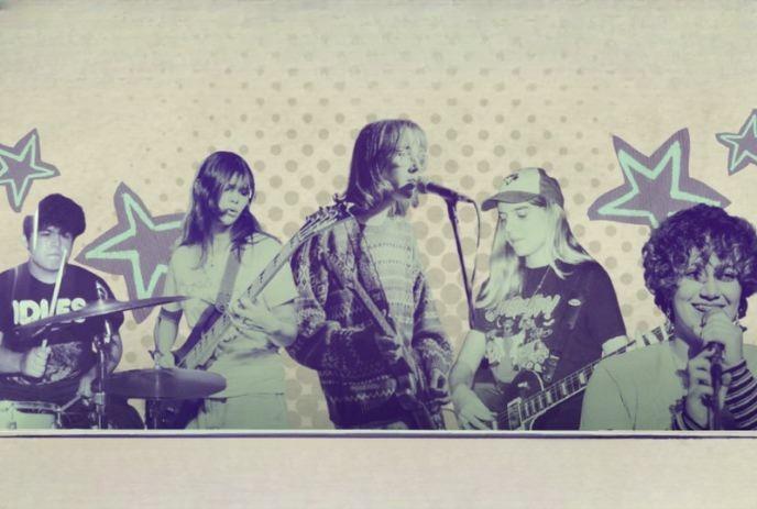 Five musicians perform together with guitars bass drums and singer before bold star patterned backdrop.