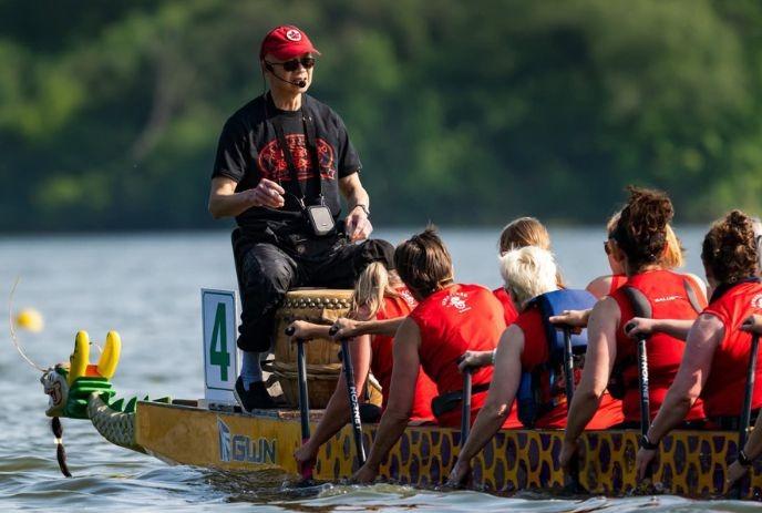 Dragon boat team in red paddles across calm water as the caller at the back guides their rhythm.