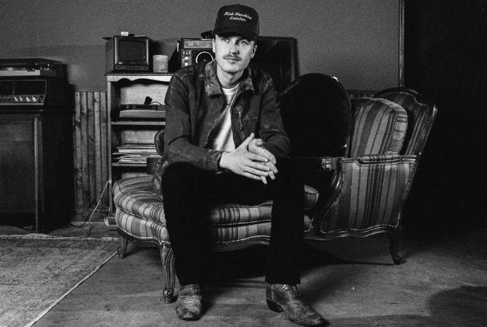 Owen Riegling sitting in a vintage room on a striped chair wearing denim jacket and hat in a black and white frame.
