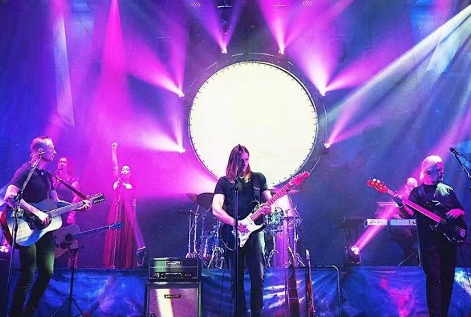 Band performing on stage under vivid purple blue lights with singers' guitarists drummer creating lively mood.