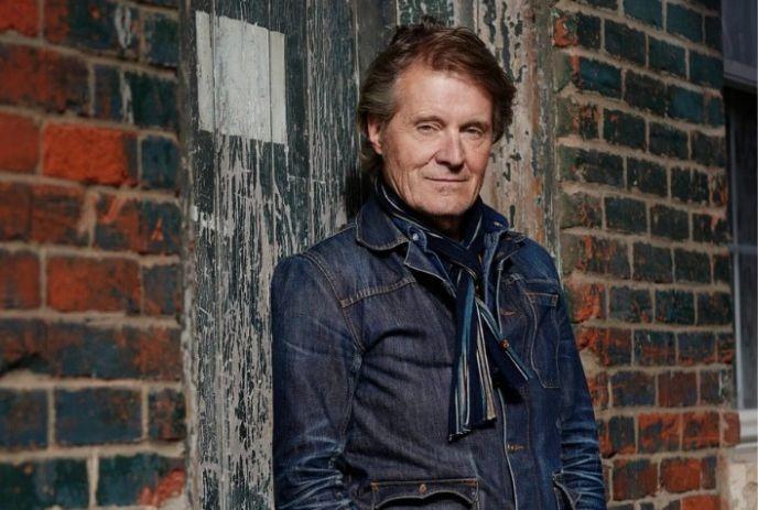 Jim Cuddy wearing a denim jacket posing for a picture under a bricked wall.