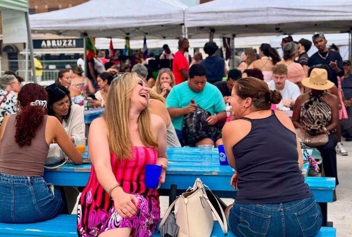 crowds gather at blue tables under white tents sharing food chatter and sunlight in a warm bustling festival.