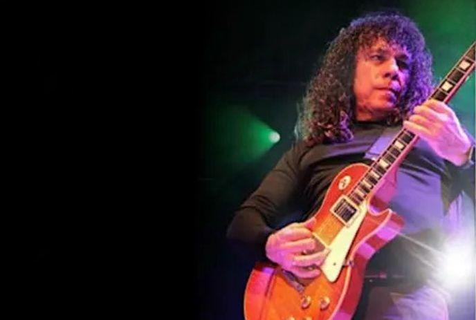 Musician with long curls plays sunburst electric guitar on dim stage lit by green light.
