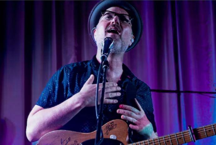 Trevor Lewington performing under purple curtains while wearing a black shirt and a hat.