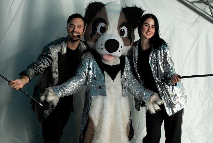Dog mascot in silver jacket stands between two performers in matching silver outfits and magic sticks.