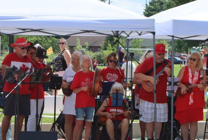 Musicians in Canada themed outfits perform outdoors beneath white tents on grass with trees around.
