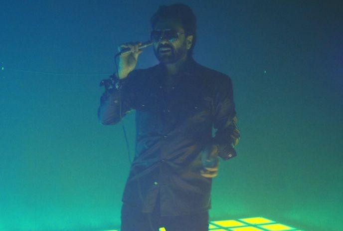 Singer in sunglasses performing on a neon-lit stage with blue and green haze.