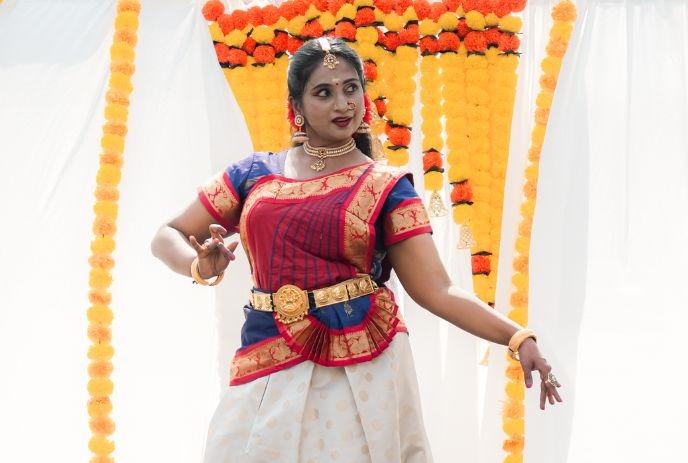 Graceful dancer in ornate red blue and gold attire posing before marigold draped backdrop in cultural performance.