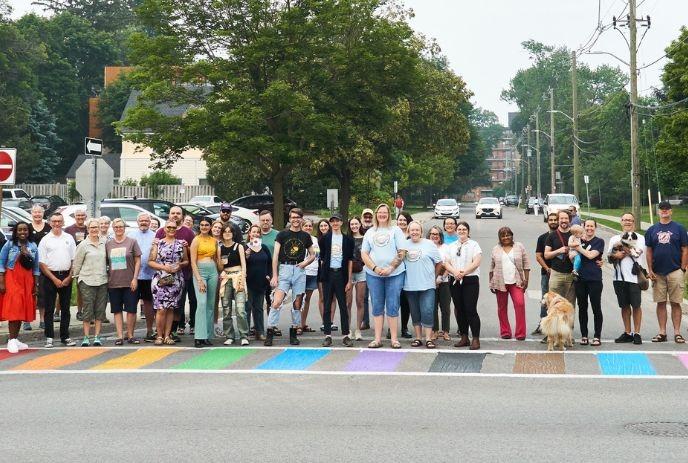 A group of people standing on the road with a rainbow zebra crossing and the green spaces in the background.