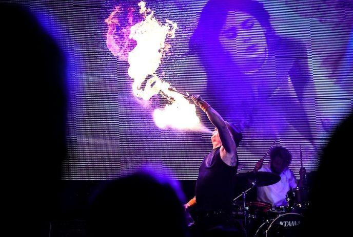 Performer ignites burst of fire onstage as drummer plays behind glowing purple and blue lights.