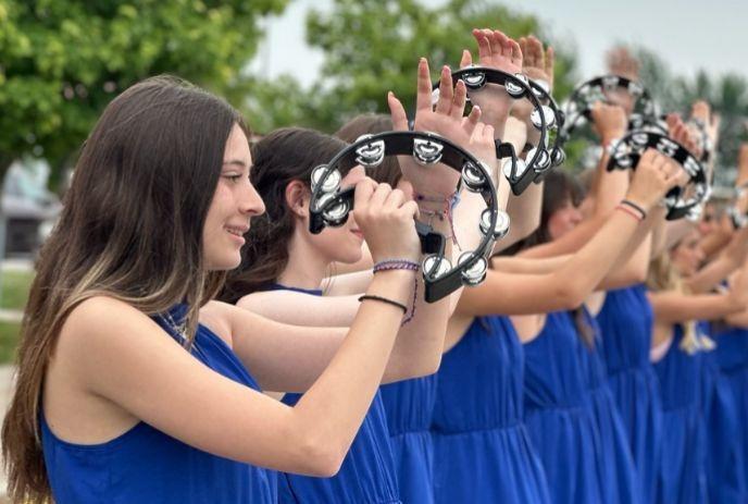 Row of performers in bright blue dresses raising tambourines outdoors with trees framing the scene.