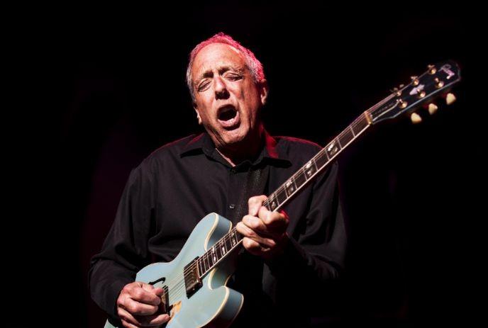 Guitarist in black plays light?blue semi?hollow guitar under dark stage lighting, fully absorbed in sound.